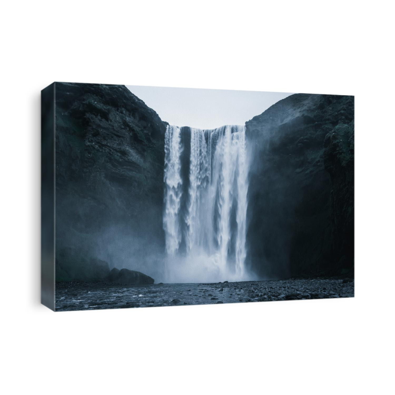 Famous Skogafoss Waterfall in Iceland from the front with rough looking water falling off the hill in a dark look with black stones in the foreground and no people in front during night